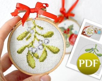May include: A hand holds a small, round, wooden-framed embroidery hoop with a cross-stitch design of a green mistletoe sprig. The hoop is decorated with a red ribbon bow. A PDF pattern for the embroidery is visible in the background.