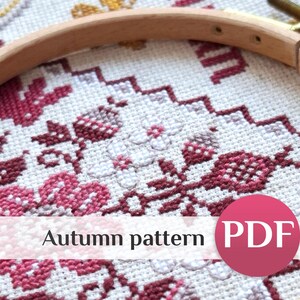 May include: A wooden embroidery hoop framing a cross-stitch project on white fabric. The design features floral and botanical motifs in shades of deep red, pink, and light purple. A white horizontal banner displays the text Autumn pattern PDF.