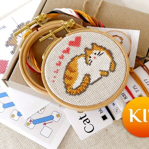 May include: A cross-stitch kit featuring a cat design with orange and white fur, surrounded by pink hearts. The kit includes embroidery hoops, thread, and instructions. The word "KIT" is printed on a round orange sticker.