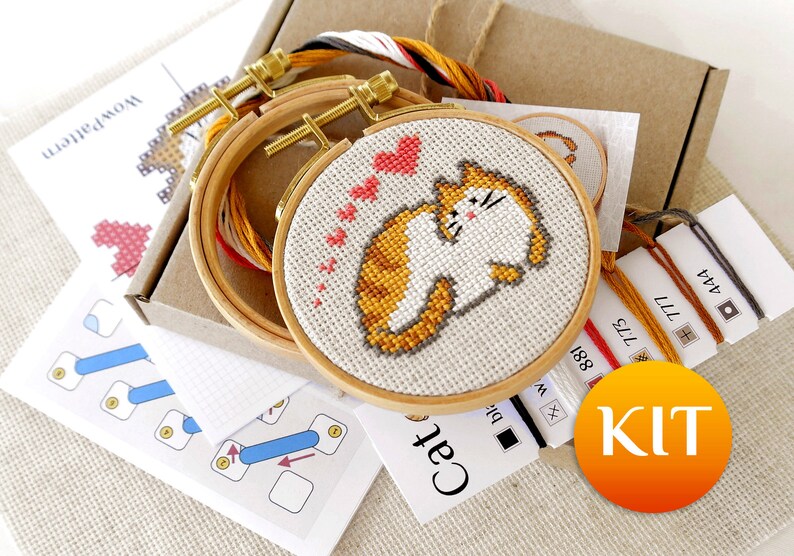 May include: A cross-stitch kit featuring a cat design with orange and white fur, surrounded by pink hearts. The kit includes embroidery hoops, thread, and instructions. The word "KIT" is printed on a round orange sticker.