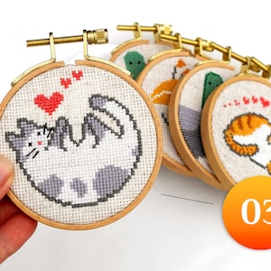 May include: A collection of small, round cross-stitch hoops with cat designs. Each hoop features a different cat illustration in various colors, including gray, orange, and white, with red heart accents. The hoops are held together by a gold-colored metal bar.