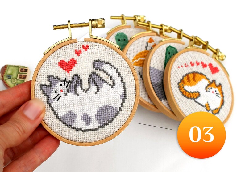May include: A collection of small, round cross-stitch hoops with cat designs. Each hoop features a different cat illustration in various colors, including gray, orange, and white, with red heart accents. The hoops are held together by a gold-colored metal bar.