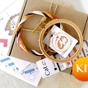 May include: A cross-stitch kit featuring a cartoon cat design. The kit includes a wooden embroidery hoop, colorful thread, a needle, and instructions. The kit is packaged in a brown cardboard box and includes a small embroidery hoop with a completed cat design.