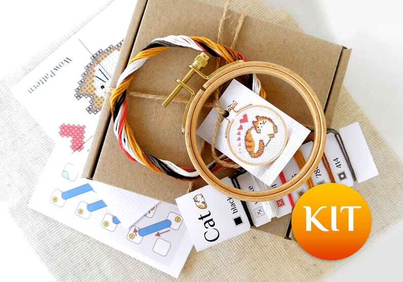 May include: A cross-stitch kit featuring a cartoon cat design. The kit includes a wooden embroidery hoop, colorful thread, a needle, and instructions. The kit is packaged in a brown cardboard box and includes a small embroidery hoop with a completed cat design.