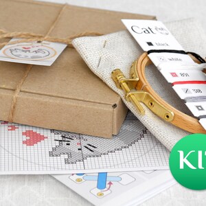 May include: A cross-stitch kit with a cat design. The kit includes a wooden embroidery hoop, fabric, embroidery floss in various colors, and a pattern. The kit is packaged in a brown cardboard box tied with twine.