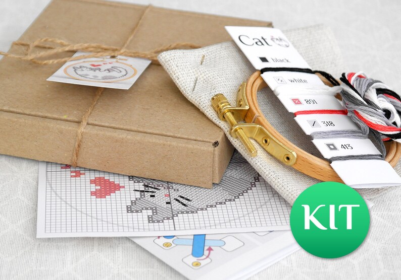 May include: A cross-stitch kit with a cat design. The kit includes a wooden embroidery hoop, fabric, embroidery floss in various colors, and a pattern. The kit is packaged in a brown cardboard box tied with twine.