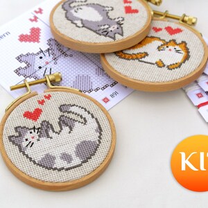 May include: Cross-stitch kits featuring playful cat designs in wooden hoops. The kits include pre-printed patterns, embroidery floss, and needles. The cats are depicted in various poses with hearts, perfect for a DIY craft project.