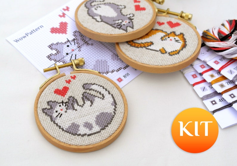 May include: Cross-stitch kits featuring playful cat designs in wooden hoops. The kits include pre-printed patterns, embroidery floss, and needles. The cats are depicted in various poses with hearts, perfect for a DIY craft project.
