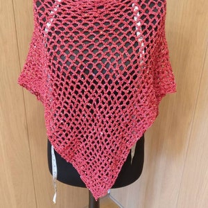May include: A red crocheted poncho displayed on a black mannequin. The poncho has a high, open neckline and a diamond-shaped open weave pattern. The garment is made of a soft, textured yarn and is designed to be worn over the shoulders.