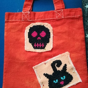 May include: A red canvas tote bag with two cross-stitch designs. The first design is a black skull with pink eyes and teeth. The second design is a black cat with blue eyes and a pink nose.