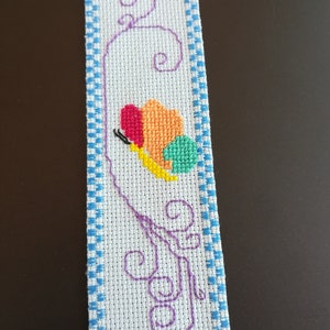 May include: A white bookmark with a blue and white checked border. A colourful butterfly is embroidered in the centre with a purple vine design.