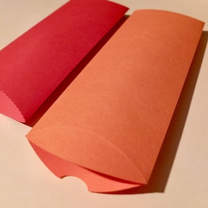 Può includere: Sono esposte due scatole di carta a forma di cuscino, una rossa e una arancione. Le scatole hanno estremità curve e sono progettate per contenere piccoli oggetti. Le scatole sono realizzate in carta strutturata.