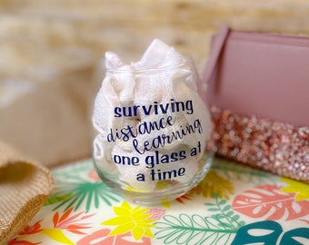 May include: A clear glass with the text "surviving distance learning one glass at a time" in blue lettering. The glass is sitting on a colorful tropical patterned surface.