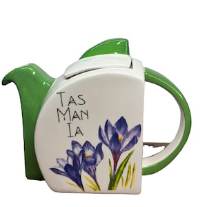 May include: White ceramic teapot with a green lid and handle. The teapot features a floral design with purple crocuses and green leaves. The text "Tas Man : Ia" is printed on the front of the teapot.