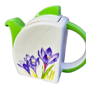 May include: A white ceramic teapot with a green lid and handle. The teapot is decorated with purple crocuses and green leaves.