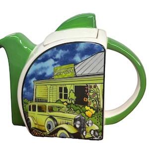 May include: Green ceramic teapot with a white lid and handle. The teapot is decorated with a scene of a yellow car parked in front of a green building with the word "Harvest" on the sign. The building has a white roof and a blue sky with clouds in the background.