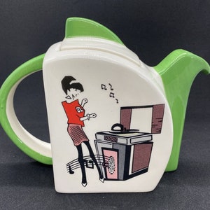 May include: A white ceramic teapot with a green lid and handle. The teapot features a retro illustration of a woman dancing in front of a record player with music notes.