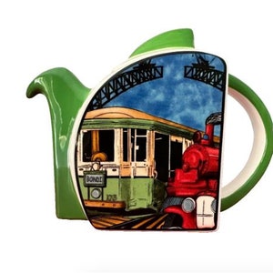 May include: Green ceramic teapot featuring a colourful illustration of a red train and a green tram with the word "BONDI" on the side. The teapot has a white handle and spout.