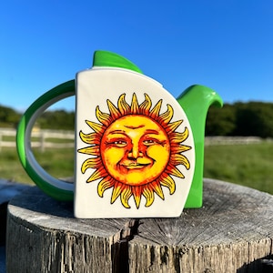 May include: A green and white ceramic teapot featuring a smiling sun face design. The sun has a yellow face with orange hair and a red mouth.