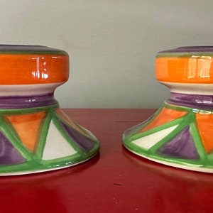 May include: Two ceramic candle holders with a geometric design. The holders are white with orange, green, and purple accents.