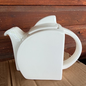 May include: A glossy white ceramic teapot featuring a distinct geometric silhouette with a flat, vertical front panel, a curved handle, a short spout, and a matching lid with a prominent, fin-like handle on top, showcasing a smooth, solid finish.
