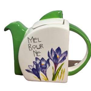 May include: White ceramic teapot with a green lid and handle. The teapot features a floral design with purple crocuses and the text "MELBOURNE" on the front.