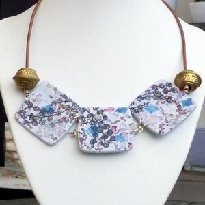 May include: A unique necklace featuring three square pendants with a watercolor-like design in shades of blue, purple, and white. The pendants are connected by silver-toned links and hang from a brown leather cord. The necklace is accented with gold-colored beads.