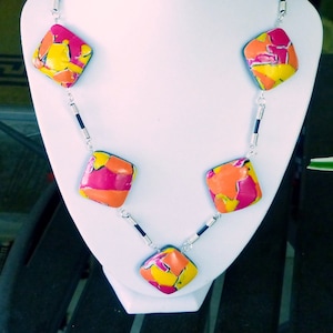 May include: A necklace with five colorful, square-shaped pendants. The pendants are made of a pink, orange, and yellow marbled material. The necklace is on a silver chain with black accents.