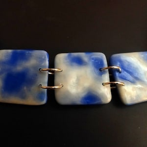 Peut inclure: Un collier en marbre bleu et blanc avec des accents argentés et dorés. Le collier est composé de trois pièces carrées reliées par des anneaux argentés.