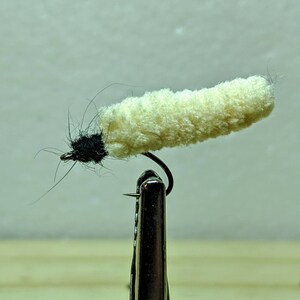 May include: A white and black fly fishing lure with a fluffy white body and a black head. The lure is attached to a hook and is held in a vise.
