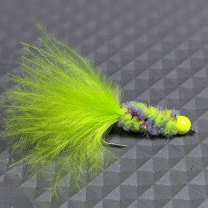 May include: A green and purple fly fishing lure with a yellow bead. The lure has a long, fluffy tail.