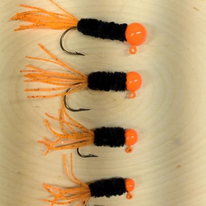 May include: Four fishing jigs with black bodies, orange tails, and orange ball-shaped heads. The hooks are black and curved. The jigs are arranged in a vertical line against a light wood grain background.