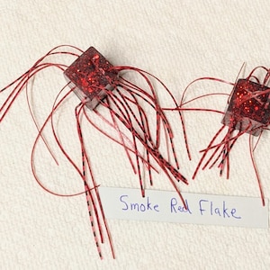 May include: Two red and black square-shaped fishing lures with long red and black strands of material attached to them. The text "Smoke Red Flake" is written on a white piece of paper below the lures.