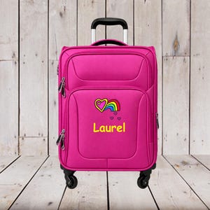 May include: A bright pink rolling suitcase with a retractable black handle and four wheels. The front features an embroidered design of a yellow heart with a rainbow and the name Laurel in yellow text, accompanied by three small floating hearts.