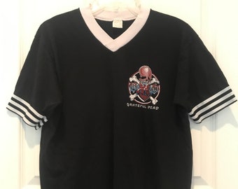May include: Black short-sleeved t-shirt with a white v-neck collar and white stripes on the sleeves. The shirt features a graphic of a skull and crossbones with a heart and the text "Grateful Dead".