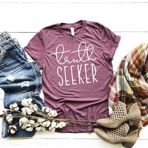 May include: A maroon t-shirt with the text "truth seeker" in white lettering. The shirt is laid flat on a white wooden surface with a pair of blue jeans, a plaid scarf, and a cotton branch.