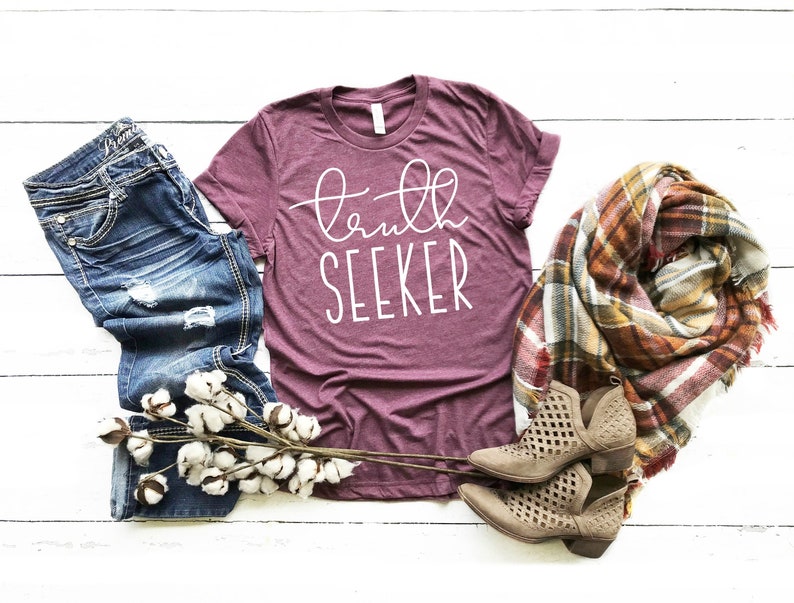 May include: A maroon t-shirt with the text "truth seeker" in white lettering. The shirt is laid flat on a white wooden surface with a pair of blue jeans, a plaid scarf, and a cotton branch.