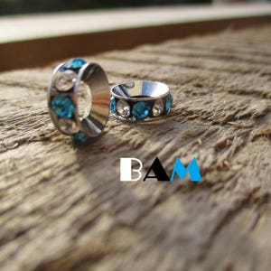 May include: Two silver rings with blue and clear rhinestones. The rings are on a wooden surface.