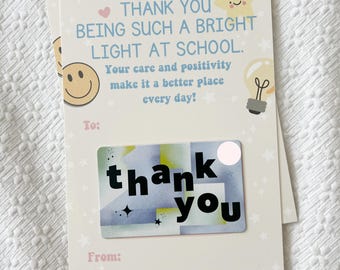 May include: A white card with a blue and green gift card that says "thank you" on it. The card has a pink heart, a yellow star, and a lightbulb on it. The text on the card says "Thank you being such a bright light at school. Your care and positivity make it a better place every day!"