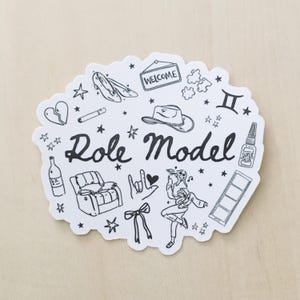 May include: A white sticker with black line art illustrations of various objects, including a cowboy hat, high heels, a bottle, and a dancer. The words "Role Model" are written in cursive.
