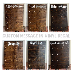 May include: Six wooden boards with a dark brown stain and white vinyl lettering. The boards have holes drilled into them and are designed to hold donuts, bagels, or other treats. The boards are decorated with various phrases, including "A Hole Lotta Love", "Treat Yourself", "Hole In One", "Donuts", "Bagel Bar", and "Donut mind if I do!"
