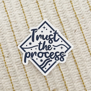 May include: White diamond-shaped sticker with the text "Trust the process" in navy blue lettering. The sticker has a navy blue border and stars and wavy lines.