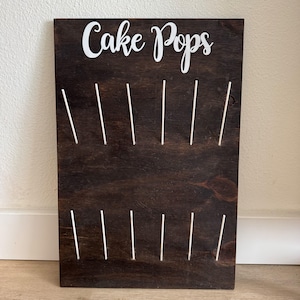 May include: A dark brown wooden cake pop display board with white text that reads "Cake Pops". The board has 12 holes for holding cake pops. The board is rectangular and has a smooth finish.
