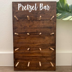 May include: A dark brown wooden pretzel bar sign with the words "Pretzel Bar" in white script. The sign has multiple wooden pegs for holding pretzels. The sign is rectangular and has a smooth finish.