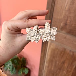 Può includere: Un paio di orecchini a fiore in perline color crema. Ogni orecchino presenta un design floreale con più petali, realizzato con piccole perline rotonde. Gli orecchini sono tenuti su uno sfondo color pesca e una porta di legno.
