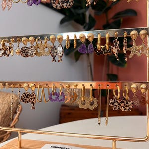 Könnte beinhalten: Ein goldfarbener Ohrring-Ausstellungsständer mit einer Vielzahl von Ohrringen. Stile umfassen Creolen, Hängeohrringe und Ohrstecker, mit Designs wie Leopardenmuster, Sternen und geometrischen Formen. Einige Ohrringe haben lila Glitzer oder florale Details.