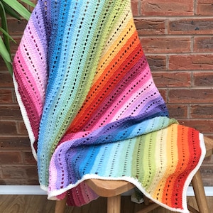 May include: A rainbow crochet blanket with a white border. The blanket is draped over a wooden chair.