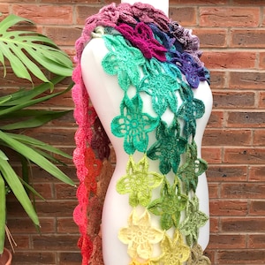 Peut inclure: Une longue écharpe en crochet colorée avec un dégradé de couleurs arc-en-ciel. L'écharpe est faite de fleurs crochetées dans une variété de nuances, du rouge foncé au jaune clair. L'écharpe est présentée sur un mannequin blanc.