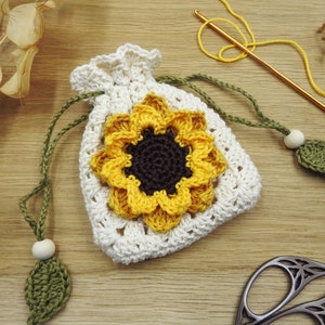 May include: A crocheted sunflower drawstring bag with a white background and yellow petals. The bag has a green crocheted leaf and a wooden bead attached to the drawstring.