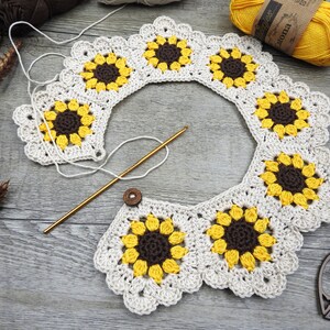 May include: A crochet project in progress, featuring a sunflower design. The piece uses cream, yellow, and brown yarn. A gold crochet hook and a wooden button are also present. The project is displayed on a grey wooden surface.